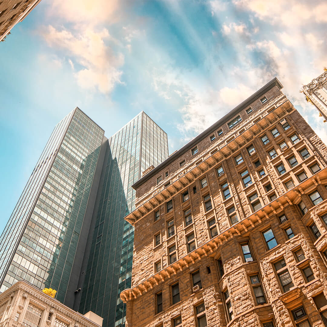 Olhando para cima em um edifício clássico de tijolos em frente a dois arranha-céus de vidro com céu azul ao fundo - Odgers Executive Search Board Headhunters